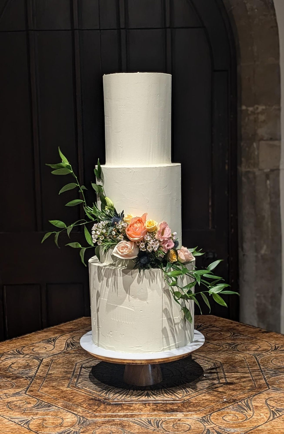 Three-tier white cake adorned with colorful flowers and leaves, set on a decorative wooden table against a dark background.