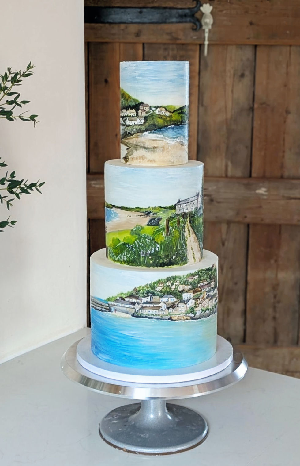 Three-tier cake adorned with detailed landscape paintings, showcasing coastal scenes. Set on a silver stand in a rustic wooden room.