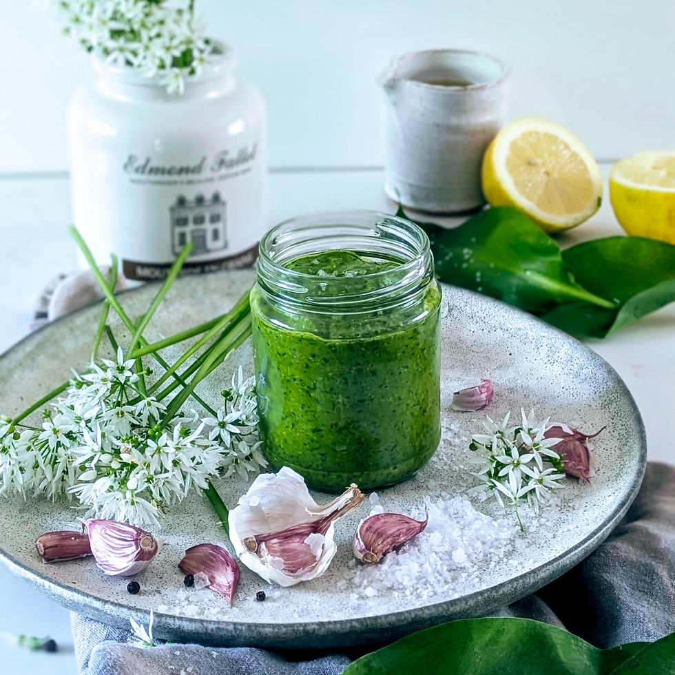 A jar of green pesto on a plate with garlic, white flowers, and salt. A lemon and ceramic items are in the background. Fresh, vibrant mood.