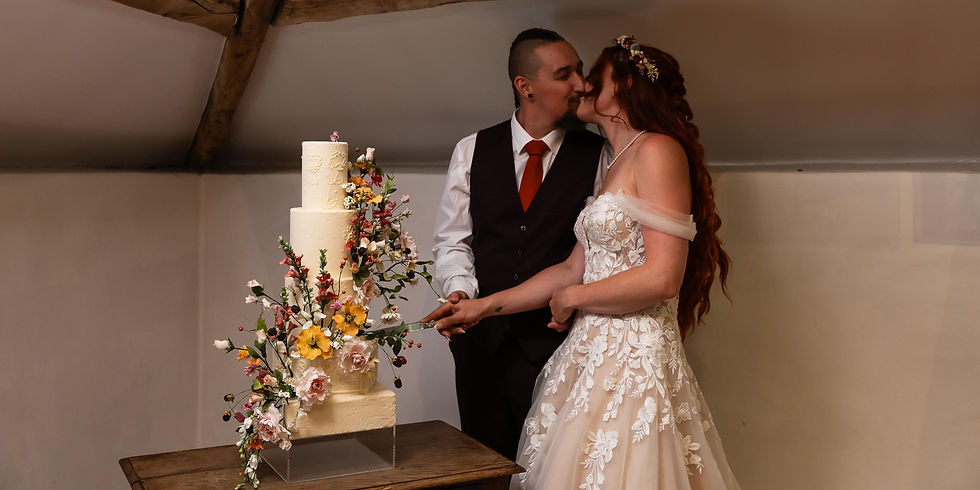 A couple cuts a floral-decorated wedding cake, sharing a kiss. She wears a lace gown; he has a vest and red tie. Cozy indoor setting.