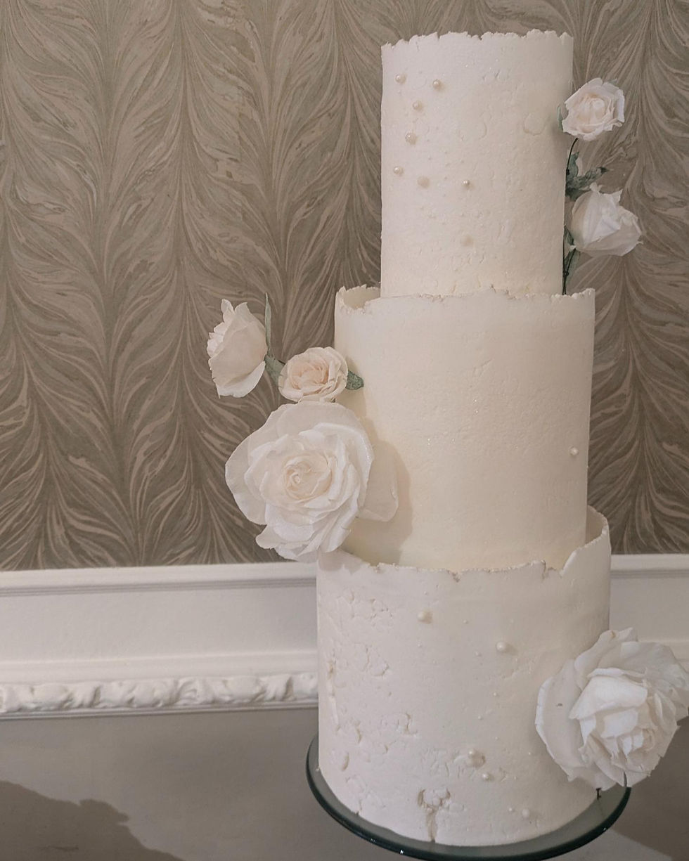 Three-tier white cake with textured icing, adorned with white roses and pearls, set against a patterned beige wall. Elegant and serene.