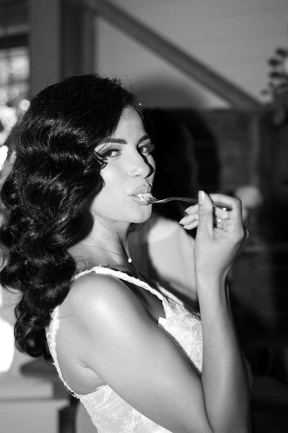 Woman in a white dress joyfully eats with a fork, smiling. Black and white image, indoor setting with blurred background.