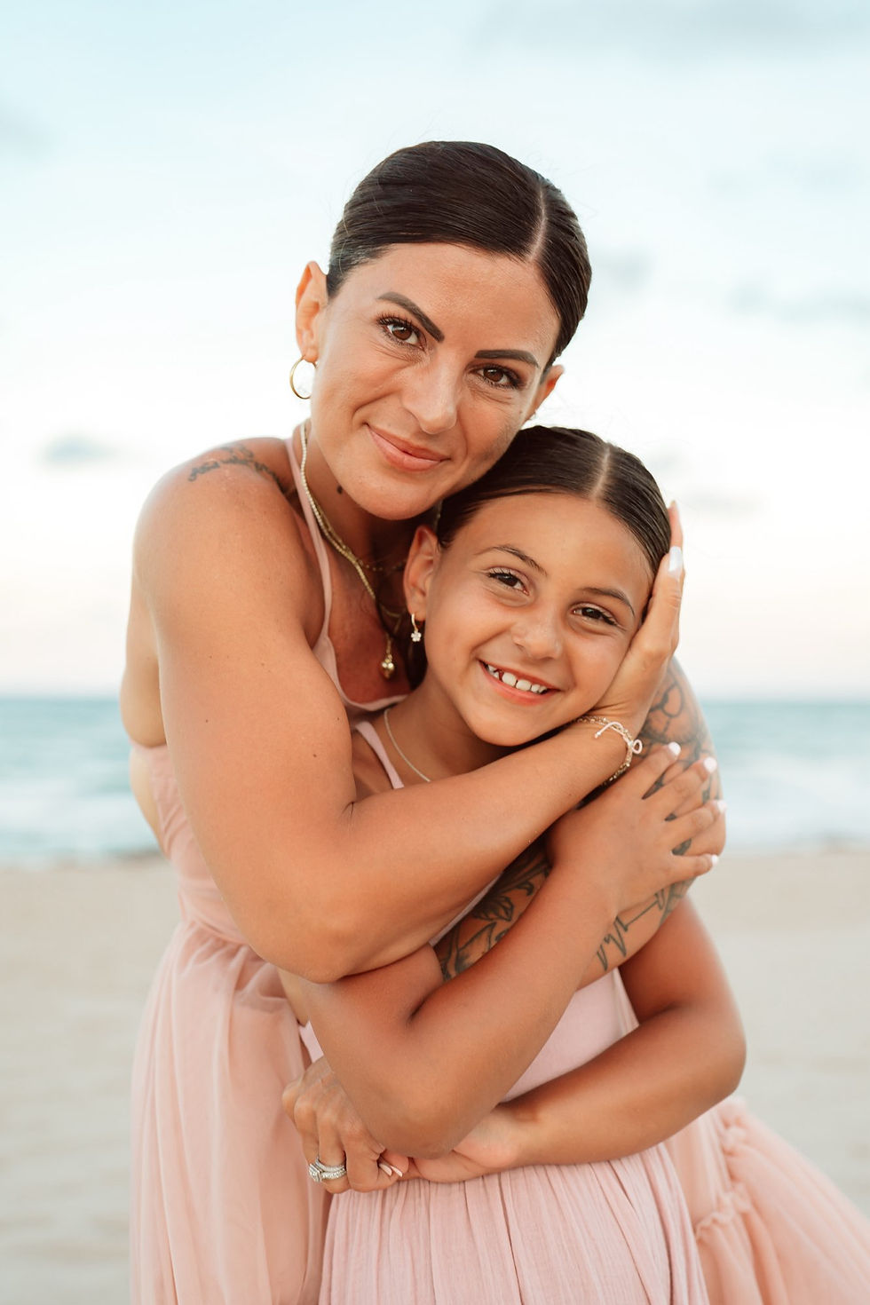 Smiling mother and daughter in pink dresses hug on a beach with calm sea and bright sky in the background.