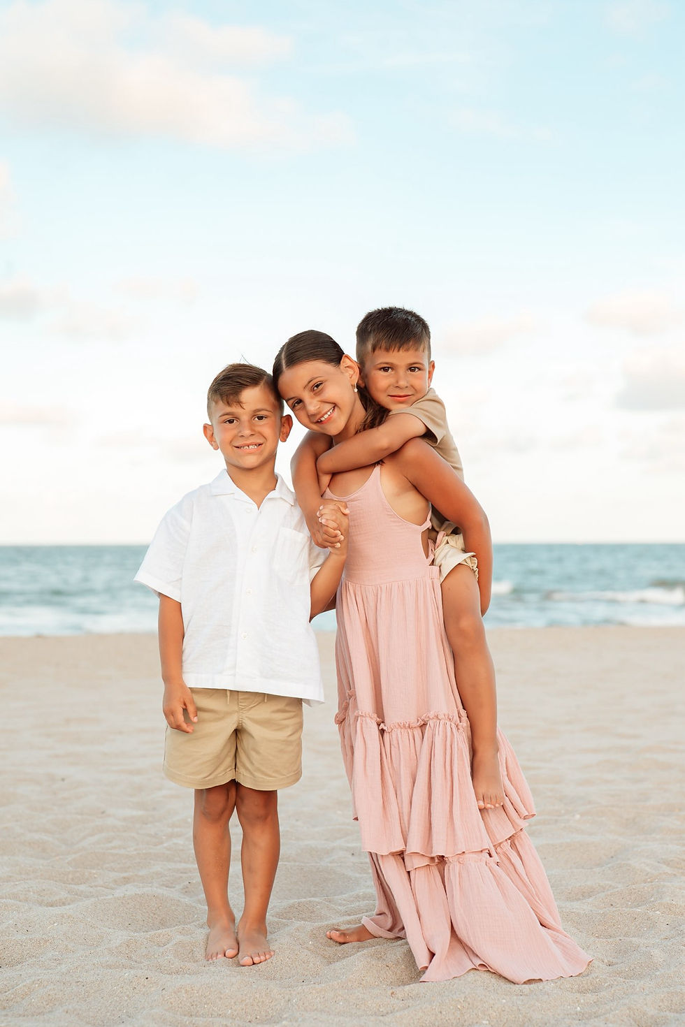 Three smiling children on a sandy beach, with a girl in a pink dress carrying a boy piggyback beside another boy in white.