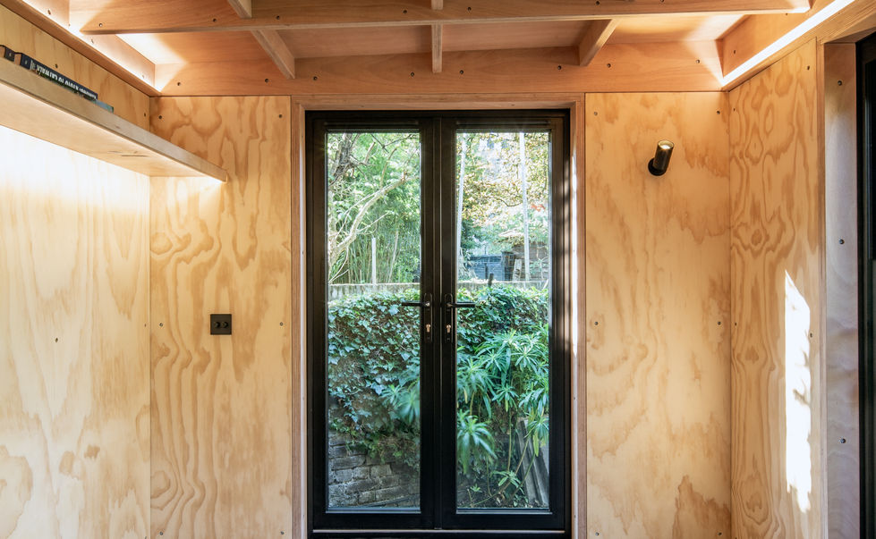 Bright interior of London garden room with plywood walls and large glass doors opening to the garden.