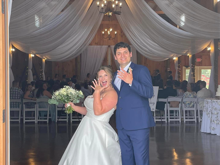 Bride and groom celebrating an elegant barn wedding venue with grand wooden doors, flowing ceiling drapery, chandeliers, and a formal reception at The Barn at Poplar Springs