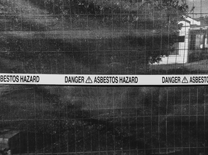 Black and white image of a fence with a caution tape reading "Danger Asbestos Hazard." Dark mesh background with partly visible buildings.