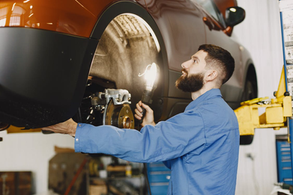 Mechanic in blue uniform examines the part of a car where the wheel goes with a work light in a garage. Brown car, focused expression, dimly lit background.
