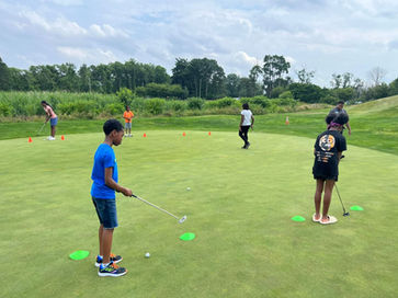 The picture depicts a group of children engaged in putting drills on a practice putting green. There are around 6 kids, ranging from around 6 to 15 years old. The children are arranged in a competition line facing each other, each taking their turn to putt a golf ball across the flat putting surface.