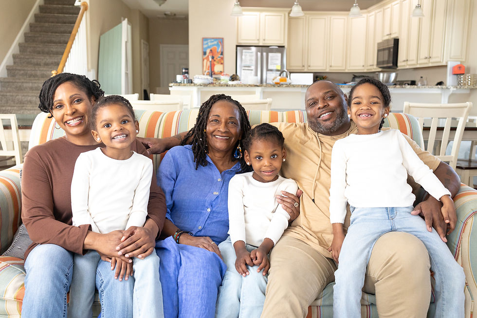 A family of six smiles on a striped couch in a bright kitchen. All wear casual outfits. The mood is joyful and relaxed.