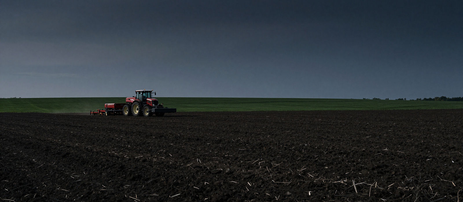 A tractor working a wide, freshly turned paddock under a dark evening sky