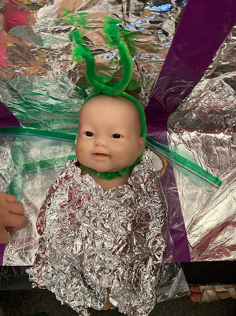 A babydoll that is wrapped in tinfoil with a green pipe cleaner wrapped around its head like an antenna.