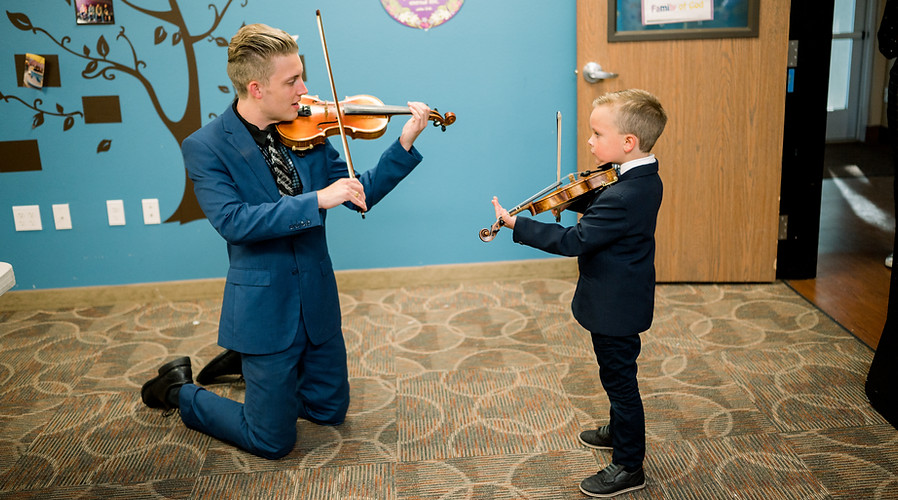 Dallin on his knees playing violin with a young boy student wearing a tux