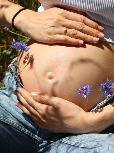 Detailaufnahme eines ungestellten Babybauchs mit blauen Wildblumen in den Jeans – Natürliche Schwangerschaftsfotografie.