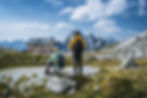 Hikers pausing on a rock and admiring a tradtional Rifugio with the Dolomite peaks on the background