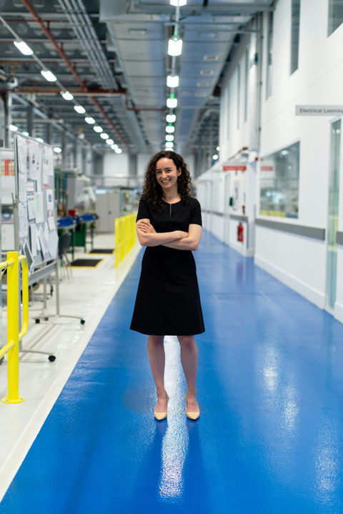 A female marketing consultant is standing in a large hall at the manufacturing site of an Australian recycling technology business. She is looking directly at the camera man who is preparing a video shoot in the production hall.