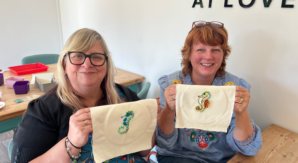 2 women smiling holding up some beaded embroidery they have made. They have both made beaded seahorses, one is green and one is gold.