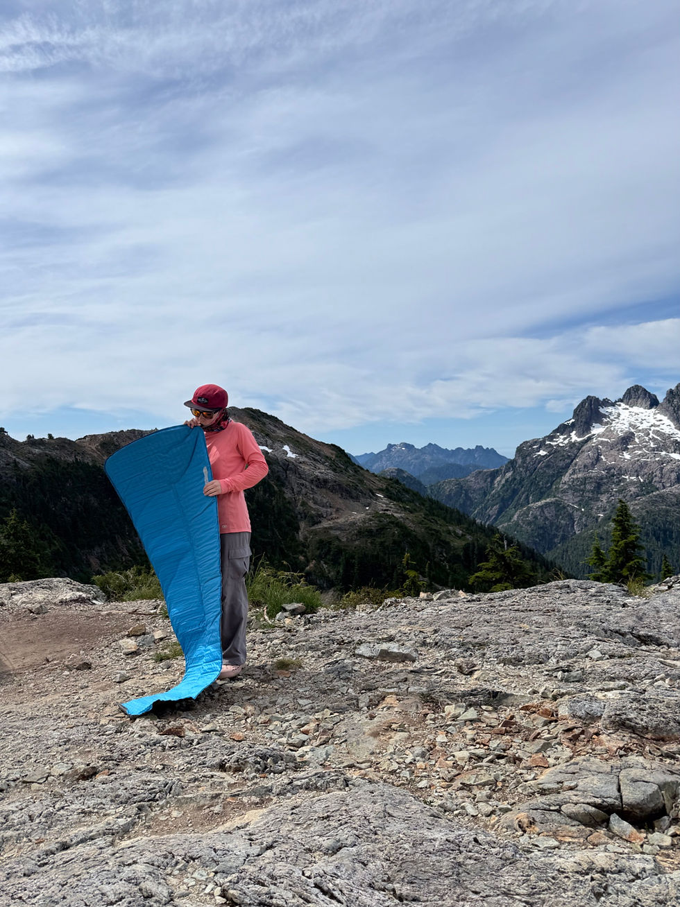 Hiker in pink jacket and red cap holds a blue sleeping pad on a rocky mountain ridge under a cloudy sky.