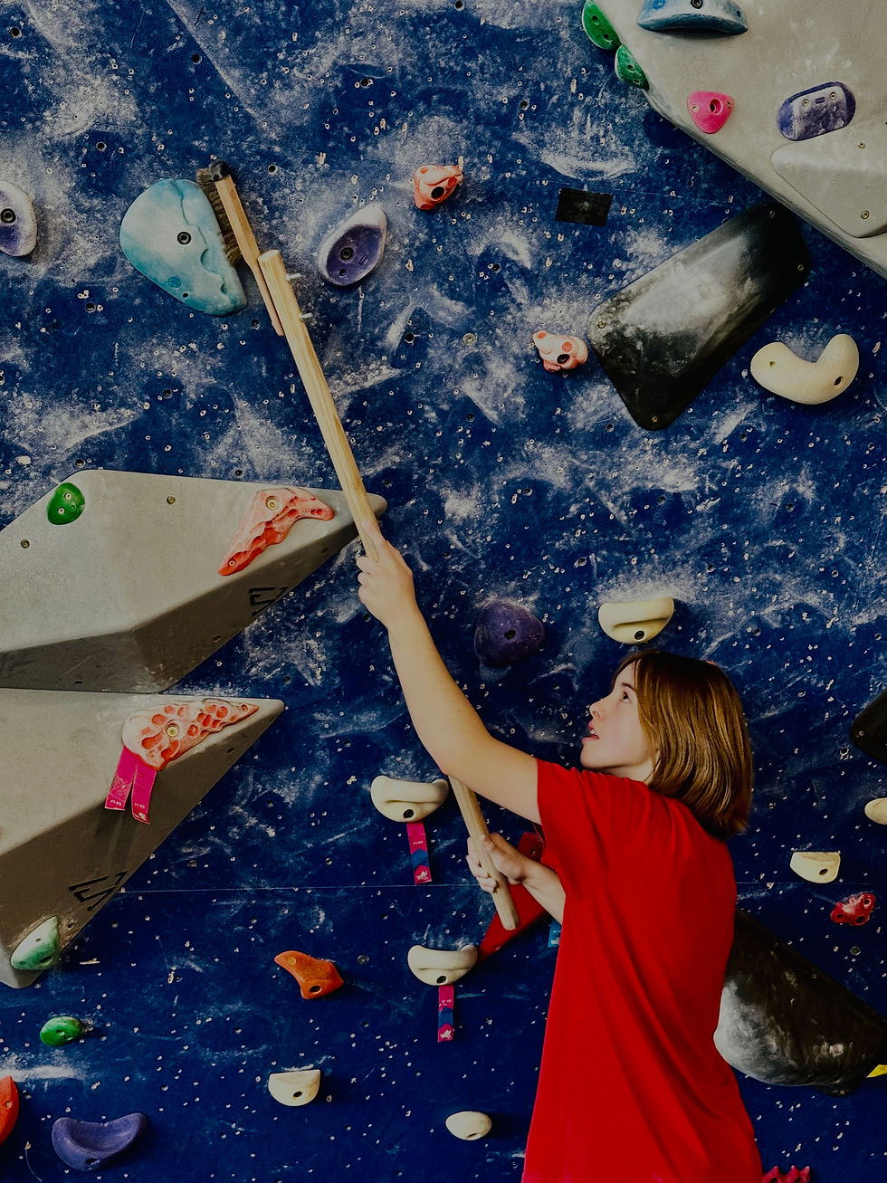 Person in red shirt brushes holds on a blue climbing wall with colorful grips, concentrating. Dust patterns create a dynamic background.