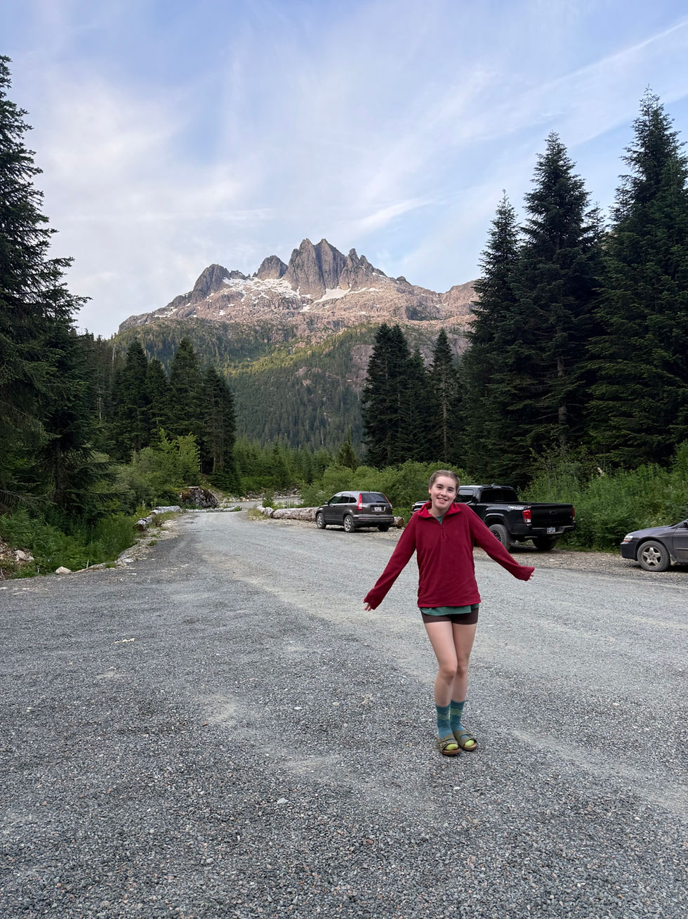Smiling girl in a red top stands on a gravel road with evergreens, parked cars, and a rugged mountain under a blue sky.