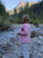 A young girl looking at sunset-lit mountains in the distance, wearing pyjamas and standing in a forest valley