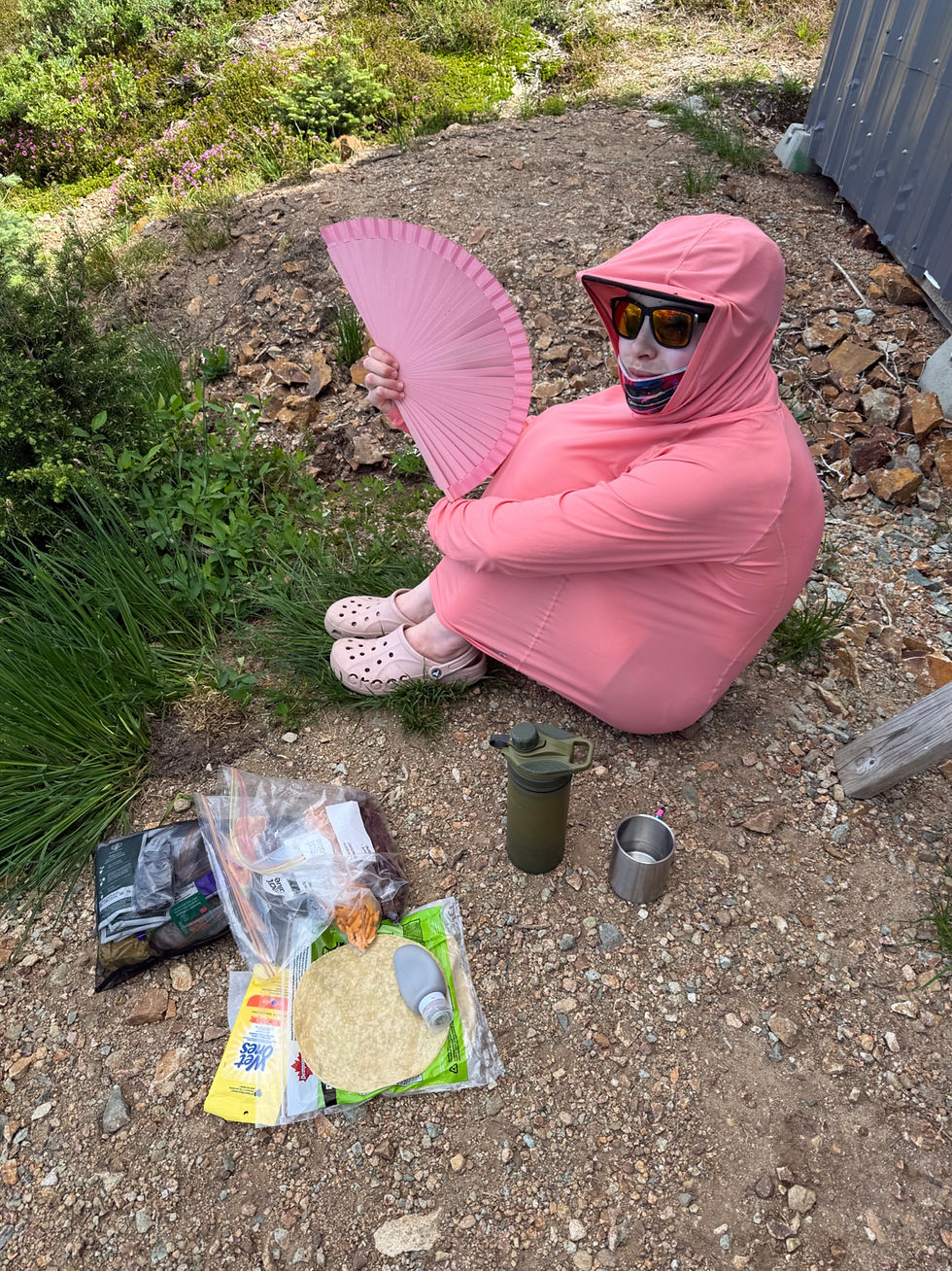 Person in pink hooded bodysuit and sunglasses sits on a rocky trail, holding a pink fan, with camping gear nearby.