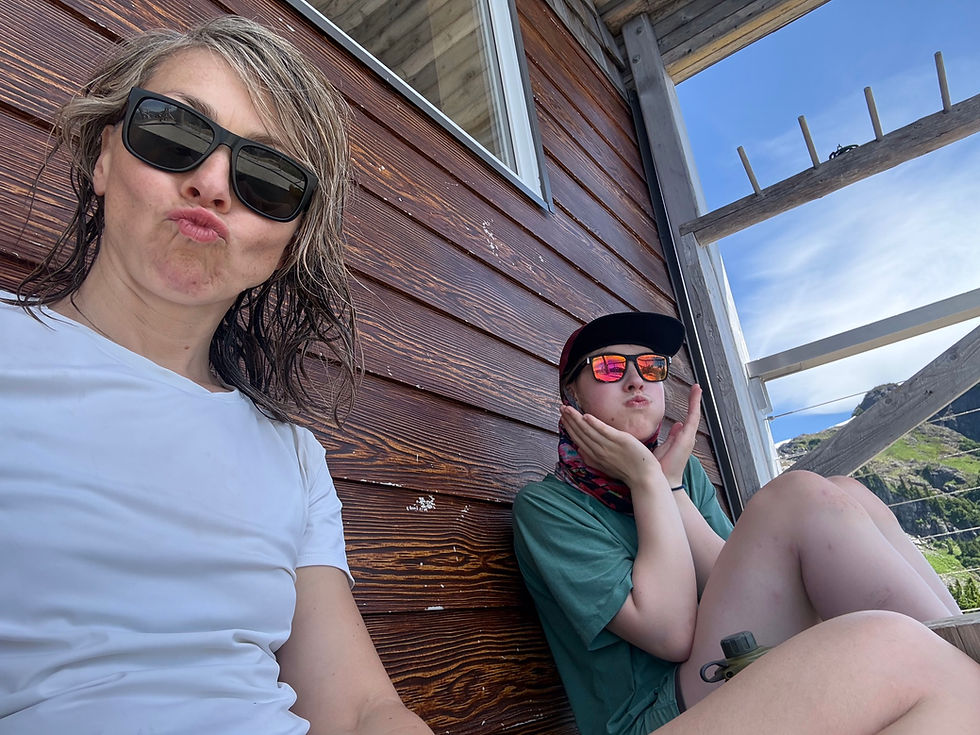 Two women in sunglasses pose on a wooden deck, one puckering lips, with blue sky and mountains behind.