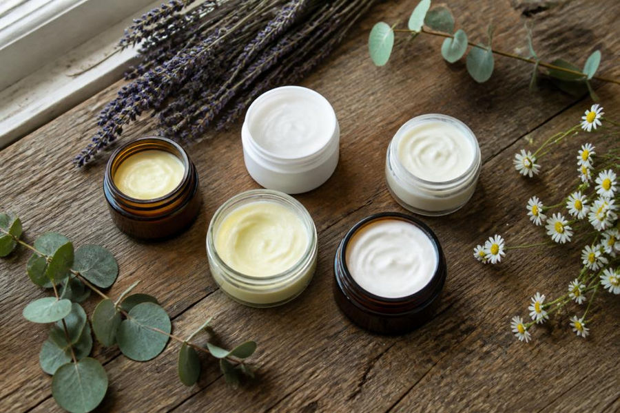 Flat lay of several small glass jars and cream containers arranged on a worn wooden surface with dried lavender sprigs, eucalyptus leaves, and small white wildflowers scattered around them, photographed in soft natural daylight from a window