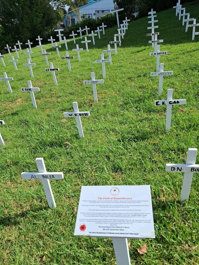 Field of white crosses on a grassy hillside memorializing fallen soldiers, with a remembrance sign in the foreground. Warkworth NZ.