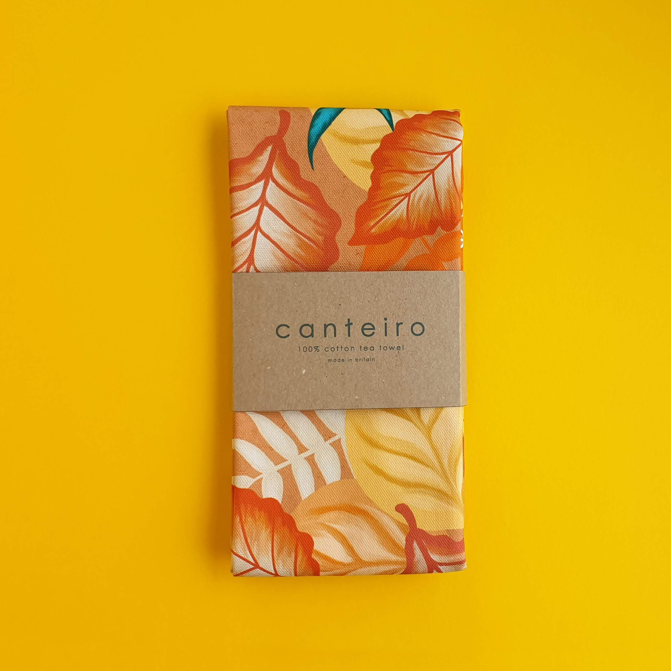 An image of a folded printed tea towel, with orange and yellow leaf print