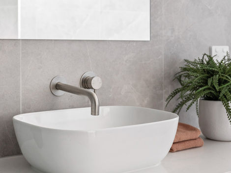White basin with wall-mounted tap on timber vanity, grey tiles and folded towel beside plant.