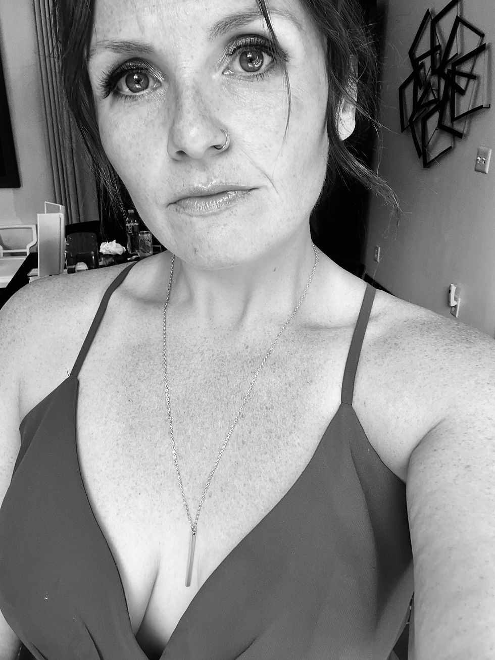 Woman with nose ring and necklace takes a black-and-white selfie, expression serious. Geometric wall art in the background.