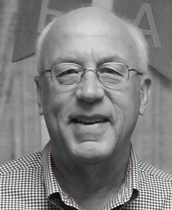 Smiling older man with glasses in a checkered shirt, neutral background. Faded banner with letters visible, grayscale filter.