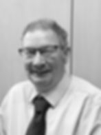 Smiling man in glasses, polka dot tie, and striped shirt in black and white photo, set against a plain wooden background.