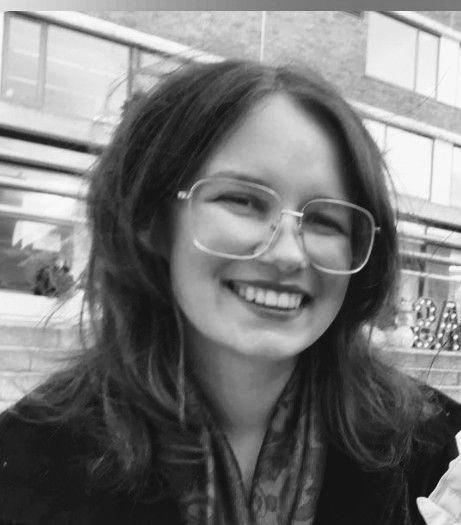 A person smiling with long hair and glasses outside a building with large windows. The black-and-white photo captures a cheerful mood.