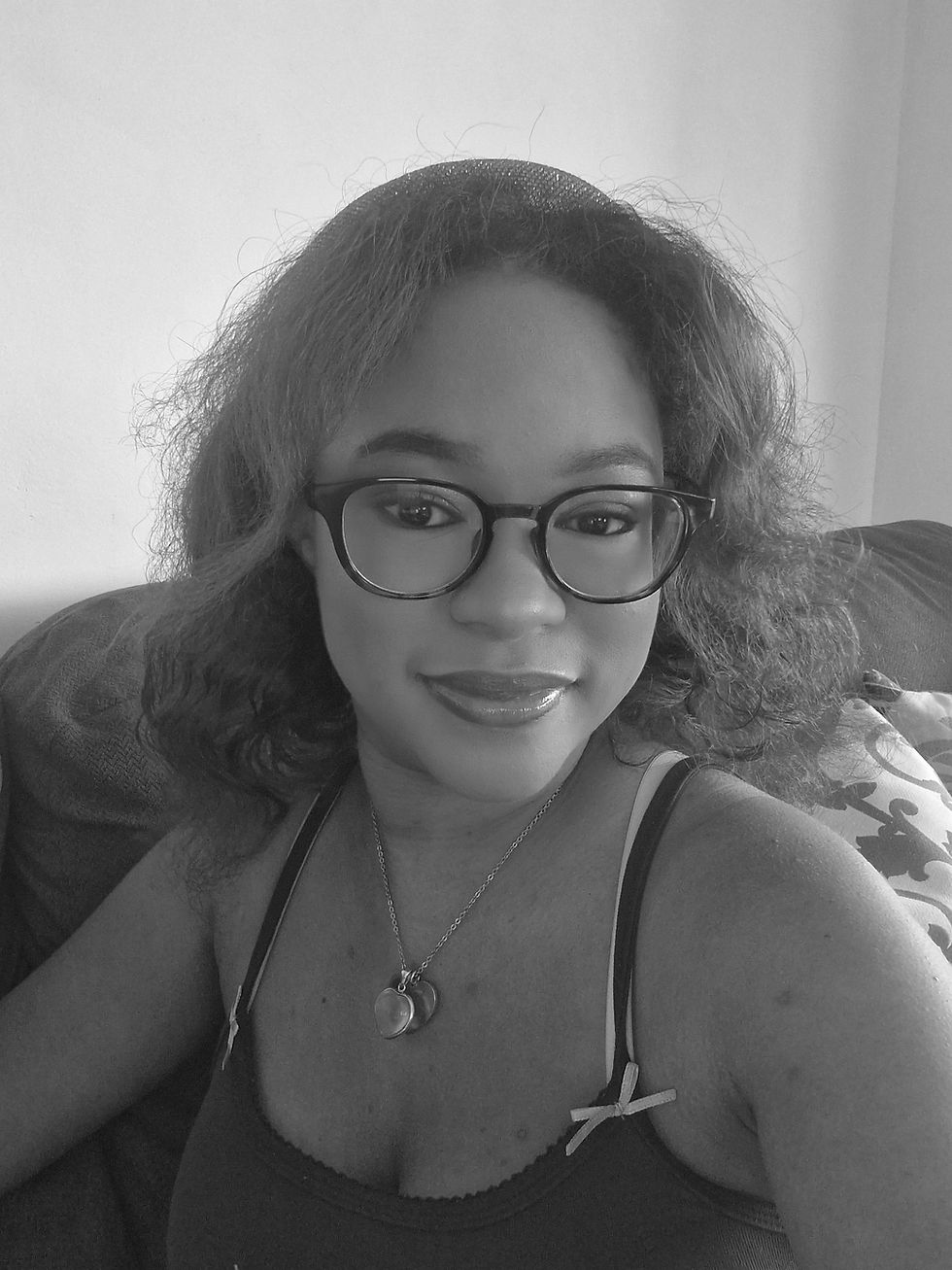 A woman with glasses and curly hair smiles at the camera, wearing a necklace and tank top. She's sitting on a couch with a cushion. Black and white.