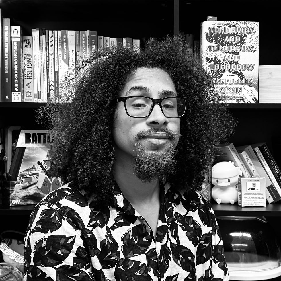 Person with curly hair and glasses, wearing a leaf-patterned shirt, smiles in front of bookshelves filled with various books and a figurine.