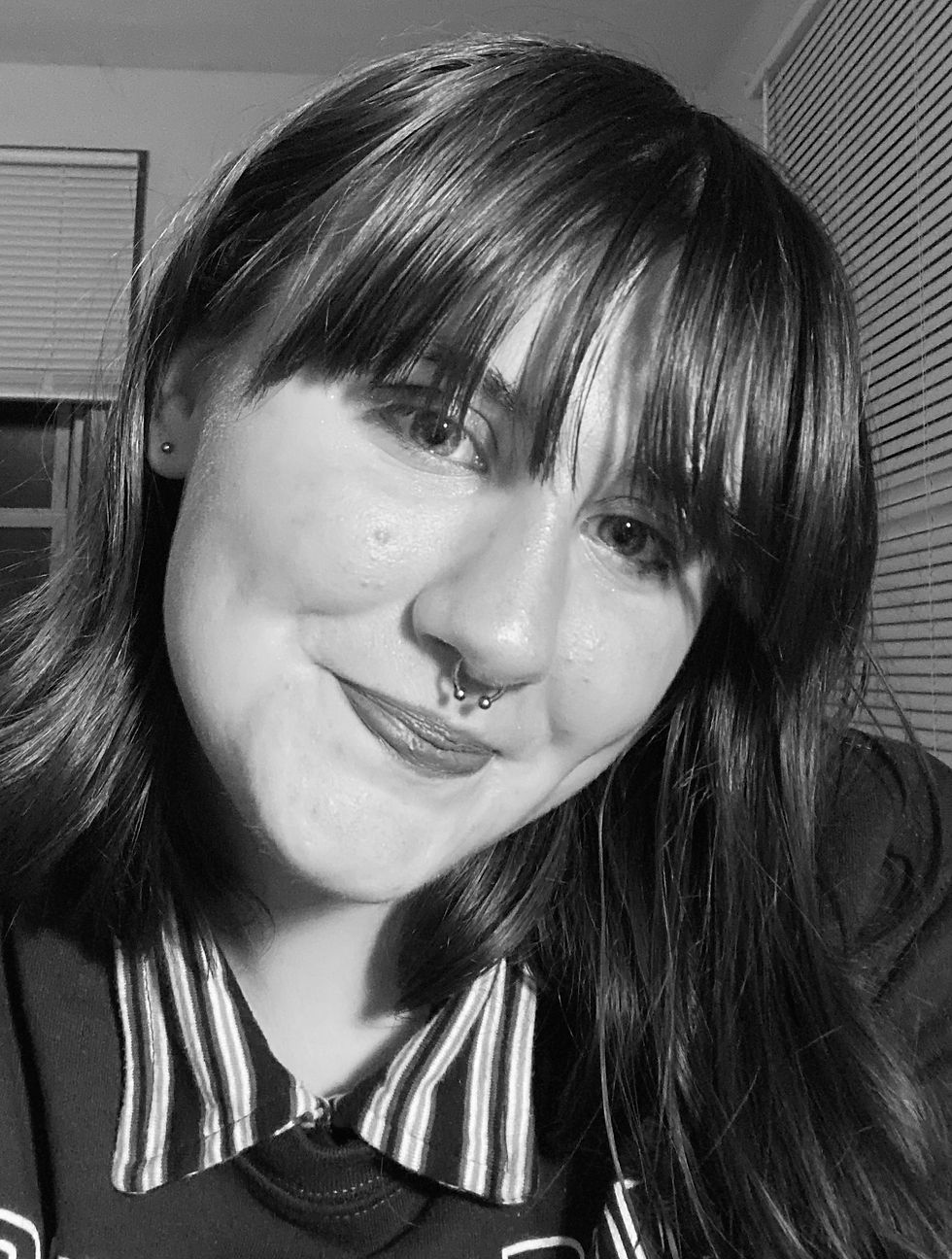 Close-up of a person with a septum piercing smiling subtly. They wear a striped-collar shirt. Blinds are visible in the background. Black and white image.
