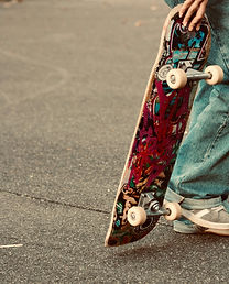 A person in jeans holds a colorful, graffiti-covered skateboard on a gray pavement, wearing white sneakers. The scene is casual and urban.