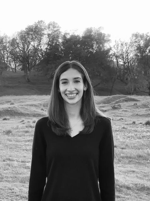 Smiling person stands in a grassy field with trees in the background. Overcast sky, black and white image, wearing a dark sweater.