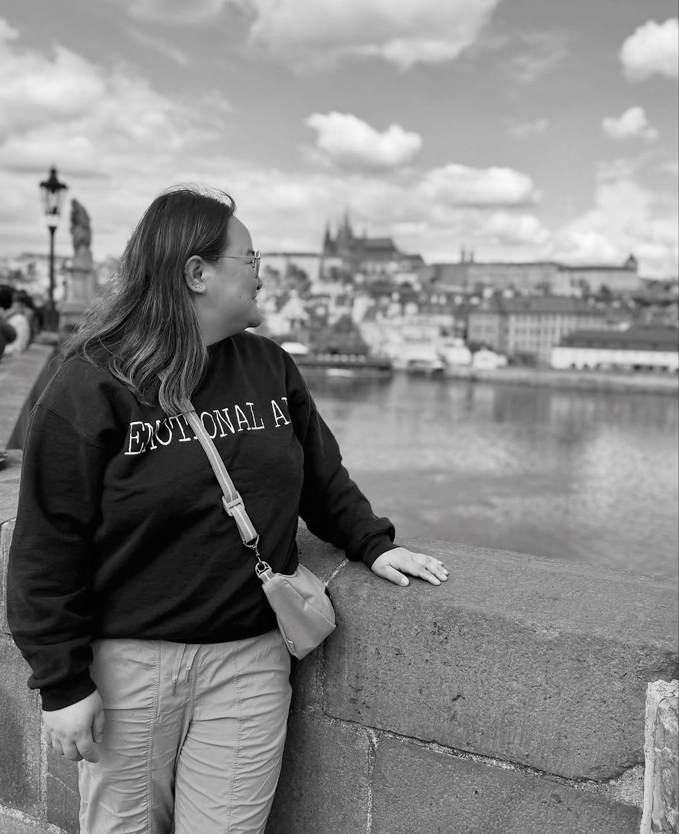 Woman in black sweater with "EMOTIONAL AS" text, leans against a stone railing, overlooking a cityscape across a river. Cloudy sky.