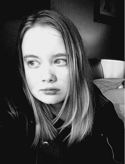 Young woman with long hair gazes thoughtfully to the side. Black and white photo with soft lighting, set in a dimly lit room.