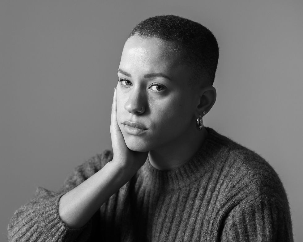 A person with short hair, wearing a sweater, rests their face on their hand, looking pensively at the camera in a grayscale portrait.