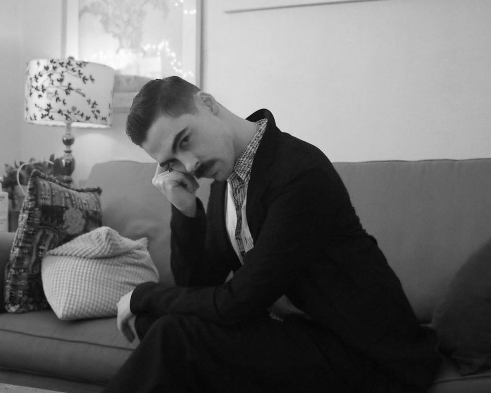 Man in a suit sits on a couch with head resting on hand, looking contemplative. Decor includes patterned pillows and a floral lamp. Black and white.
