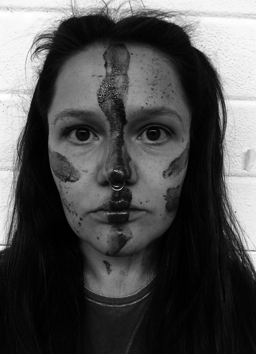 Black and white photo of a person with dark makeup streaks on their face, wearing a septum ring. White brick wall background. Intense gaze.