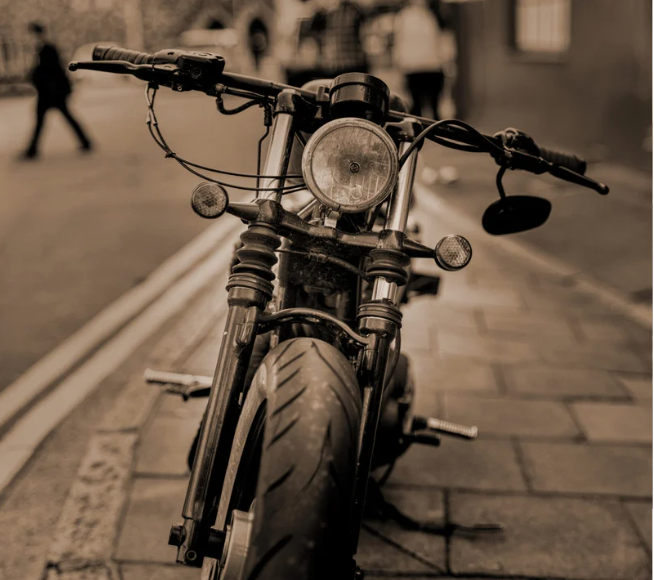 Sepia-toned image of a parked motorcycle on a cobblestone street. Blurred figures walk by in the background, creating a vintage feel.