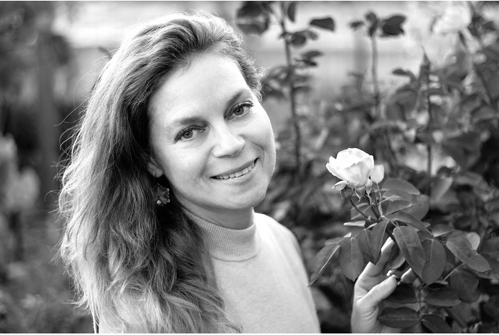 Smiling woman with long hair holds a rose in a garden. The black-and-white photo captures a serene, natural setting.