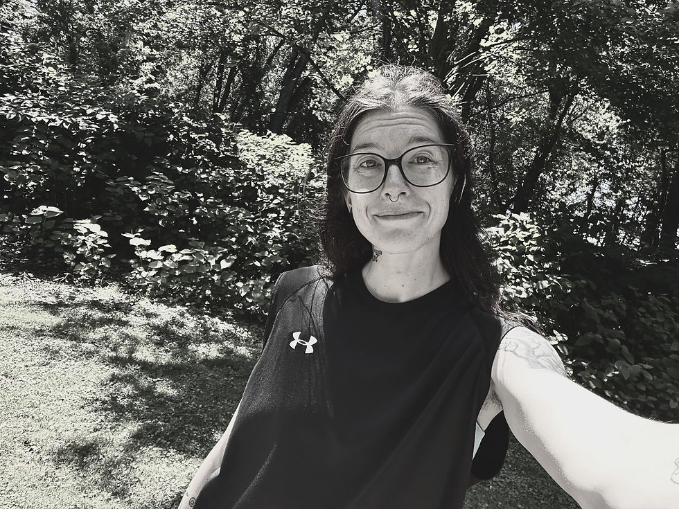 Person in glasses and dark top poses for a selfie outdoors, smiling in a wooded area. Sunlight creates dappled shadows on the ground.