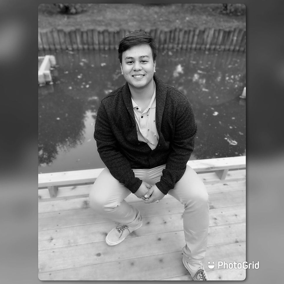 Person smiling, sitting on a wooden bench near a pond with trees in the background, wearing a dark sweater and light pants. Monochrome image.