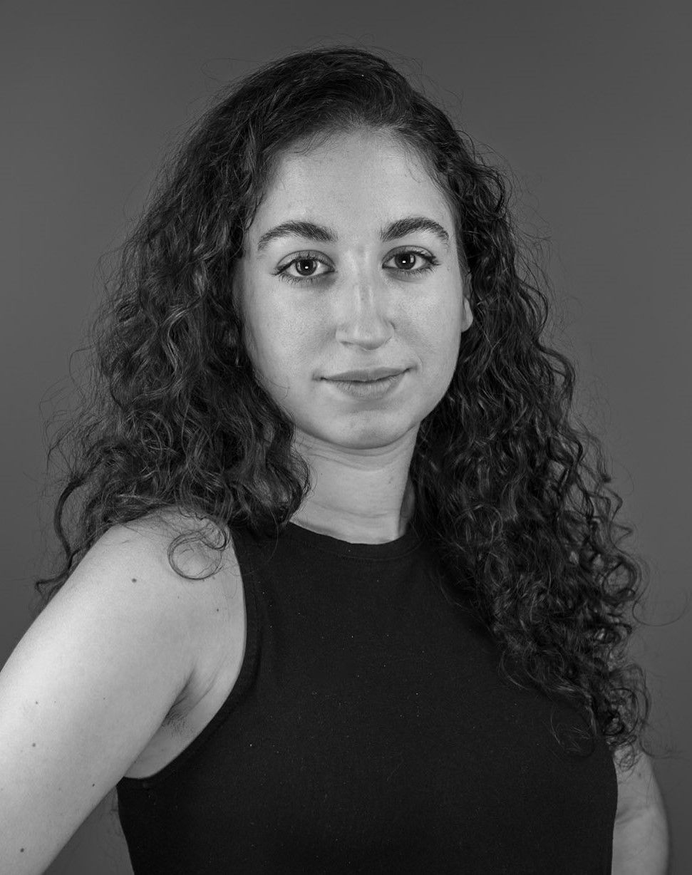 Black and white portrait of a person with curly hair in a sleeveless top, looking directly at the camera with a neutral expression. Gray background.